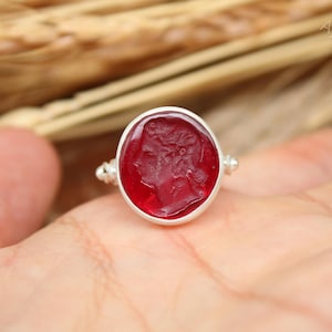 Op de afbeelding: Een zilveren ring met een grote, ovale, rode edelsteen. De edelsteen is gegraveerd met een profiel van een persoon. De ring heeft een eenvoudige zilveren band met kleine decoratieve elementen aan weerszijden van de edelsteen.