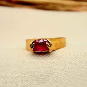 May include: A gold-colored ring with a rectangular, deep red gemstone. The ring has a simple band and the gemstone is held in place by two curved prongs. The ring is set against a cream-colored fabric.