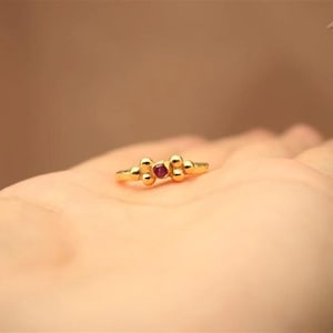 May include: A gold ring with a central red gemstone, flanked by two sets of gold beads. The ring is displayed on a person's hand. The ring is a piece of jewelry.
