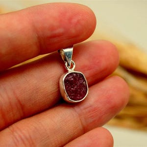 May include: A pendant featuring a small, rough-textured, deep red gemstone set in a polished silver bezel with a silver bail at the top. The text AyraSilver is visible in the upper right corner of the image. The pendant has a rounded square shape.