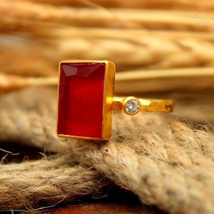 May include: A gold-colored ring with a rectangular, deep red gemstone. A small, round, clear stone is set on the band. The ring is displayed on a textured, neutral-colored surface. The ring is a piece of jewelry.