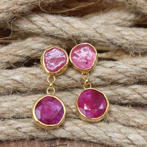 Peut inclure: Boucles d'oreilles dorées avec des pierres précieuses de couleur rubis. Chaque boucle d'oreille présente une petite pierre de forme irrégulière en haut et une pierre plus grande et ronde en dessous. Présentées sur une surface de corde brune texturée.