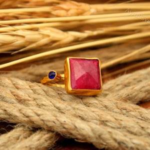 May include: A gold ring with a large, square, faceted ruby gemstone and a smaller blue gemstone. The ring is set against a backdrop of rope and wheat stalks, creating a rustic aesthetic.