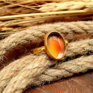 May include: A gold ring featuring an oval-cut, colour-changing stone in shades of orange, yellow, and brown. The ring sits on a thick, textured rope, with wheat stalks in the background. The ring's design is simple and elegant.