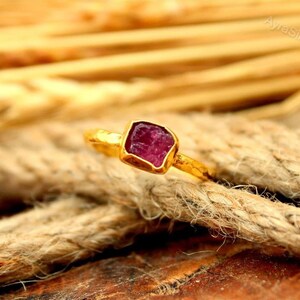May include: A gold-colored ring with a square, raw-cut ruby gemstone. The ring is set on a textured, brown surface. The ruby is a deep, rich red color. The ring's band is thin and simple.