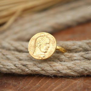May include: A gold-colored ring featuring a circular coin design with a profile of a person wearing a laurel wreath. The ring is set on a thick, textured rope, with the AyraSilver logo visible in the background.