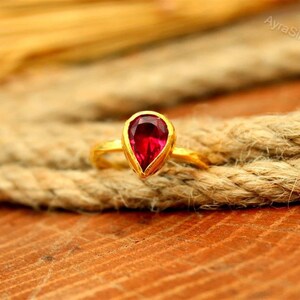 Op de afbeelding: Een gouden ring met een druppelvormige, dieprode edelsteen. De ring is geplaatst op een dik, getextureerd touw tegen een houten oppervlak. De edelsteen is het middelpunt, met de gouden band als contrasterend accent.