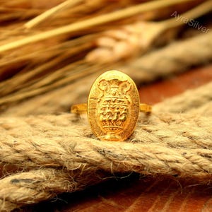 May include: A gold-toned ring featuring an oval-shaped face with an embossed design of a classical urn or vase with decorative handles and figures. The ring has a textured surface and the text AyraSilver is visible in the upper right corner.