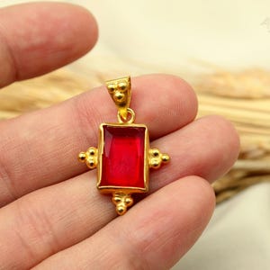 May include: A gold pendant featuring a rectangular red gemstone set in a metal frame with small decorative spheres on the sides and bottom. The pendant includes a bail at the top with three small spheres and the text AyraSilver in the corner.