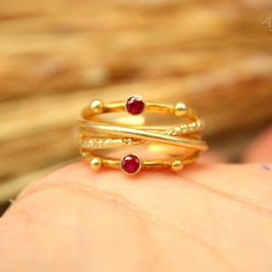 Puede incluir: Un anillo dorado con un diseño en capas, con dos pequeñas piedras preciosas rojas redondas. El anillo tiene una banda texturizada y está acentuado con pequeñas cuentas doradas redondas. Es una joya.