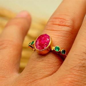 May include: A gold ring featuring a large oval ruby gemstone as the centerpiece, flanked by three smaller emeralds. The ring is set on a gold band, creating a striking contrast between the red, green, and gold colors.