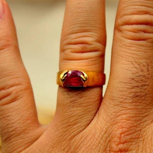 Peut inclure: Une bague en or avec une grosse pierre rouge rectangulaire sertie dans un sertissage à griffes. La bague est portée sur un doigt, mettant en valeur le design et la couleur du bijou. Le corps de la bague est doré.