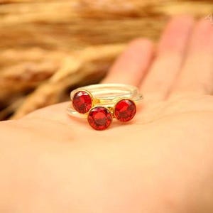 May include: A gold-toned ring with three red gemstones. The ring is set on a person's palm, with a blurred background of wheat stalks. The gemstones are round and appear to be faceted.