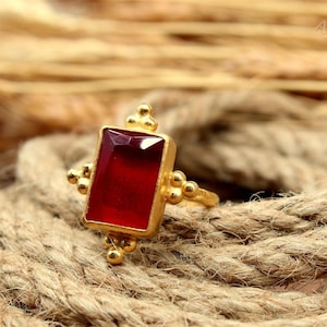 May include: A gold-colored ring with a rectangular, deep red gemstone. The ring has decorative gold accents and is set against a backdrop of natural rope. The ring is a piece of jewelry.