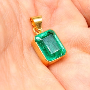 May include: A gold-colored pendant with a rectangular, emerald-cut, bright green gemstone. The pendant is held in a person's hand. The gemstone is set in a gold bezel setting.