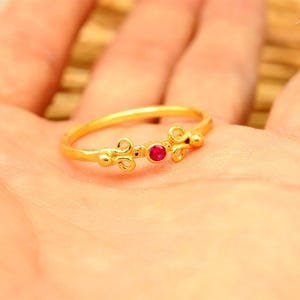 Könnte beinhalten: Ein goldfarbener Ring mit einem kleinen, runden, roten Edelstein in der Mitte. Der Ring hat ein filigranes Design mit dekorativen Elementen auf beiden Seiten des Edelsteins. Der Ring wird in einer Hand gehalten.