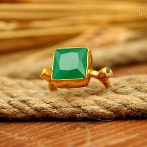 May include: A gold ring featuring a square-cut green gemstone set in a bezel, with a small clear stone accent on one side of the band. The ring has a polished finish and the text AyraSilver is visible in the upper right corner of the image.