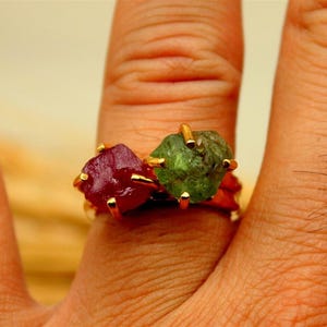May include: A gold ring featuring two raw gemstones: a deep red ruby and a green emerald. The gemstones are set side-by-side, with gold prongs securing them. The ring is displayed on a finger, showcasing the natural textures and colors of the stones.