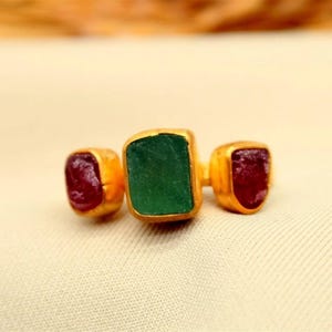 May include: A gold ring with three gemstones. The center stone is a rectangular emerald, flanked by two smaller, square-cut rubies. The ring is set on a cream-colored fabric.
