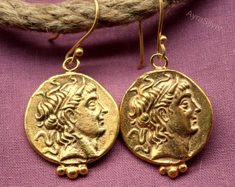 May include: A pair of gold-colored coin earrings. Each earring features a profile of a person with a laurel wreath, and a small cluster of beads at the bottom. The earrings are suspended from gold-colored hooks.