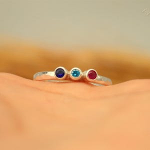 May include: A silver ring with three round gemstones in blue, teal, and red. The ring is set on a light-colored surface, with the gemstones aligned in a row. The ring is a delicate piece of jewelry.