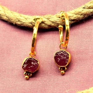 May include: Gold hoop earrings featuring a raw, deep red gemstone pendant. The earrings have a textured, organic appearance, with small gold beads at the pendant's base. The earrings are displayed on a pink surface.