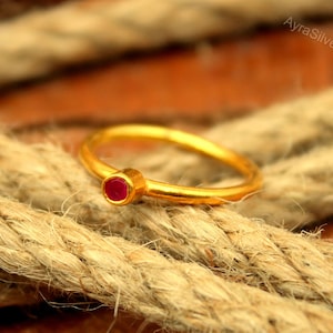 May include: A gold ring featuring a small, round, red gemstone. The ring is set on a thick, textured rope. The ring's band is thin and simple, and the gemstone is a vibrant red colour. The ring is a piece of jewellery.