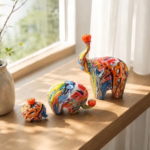 Peut inclure: Trois figurines d'éléphants arborant un motif graffiti multicolore et vibrant avec des éclaboussures de rouge, bleu, jaune et noir. Chaque éléphant est surmonté d'une petite sphère orange brillante sur la tête ou la trompe, formant un ensemble.