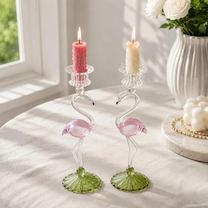 May include: Two clear glass candle holders shaped like flamingos with pink bodies and green circular bases. Each holder features a clear glass cup at the top, one containing a textured pink pillar candle and the other a textured cream pillar candle.