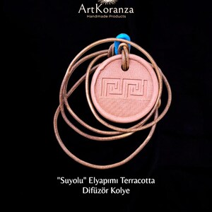 Pode incluir: Um colar com pingente de terracota com um design geométrico. O pingente é redondo e tem um cordão de couro castanho claro. O colar está sobre um fundo preto. O texto "ArtKoranza Handmade Products" está no topo.