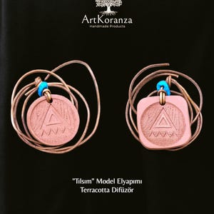 Op de afbeelding: Twee terracotta-kleurige hangers met een geometrisch ontwerp en blauwe kralen. De hangers zitten aan een bruin koord en de afbeelding bevat de tekst "Tılsım Model Elyapımı Terracotta Difüzör". Het ArtKoranza-logo staat bovenaan.