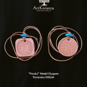 Puede incluir: Dos difusores de terracota con diseño geométrico, suspendidos por cordones beige y cuentas azules. Los difusores se muestran sobre un fondo negro. El texto "ArtKoranza Handmade Products" está en la parte superior y "Pitrak2 Model Elyapimi Terracotta Difüzör" en la inferior.