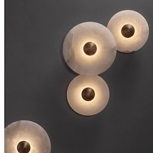 May include: Four circular wall sconces featuring translucent white stone discs with a marbled texture. Each light fixture has a central, dark metallic dome with a concentric circular pattern, emitting a soft, warm glow from behind the stone.