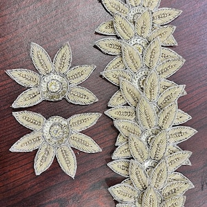 Pode incluir: Dois remendos individuais em forma de flor e uma tira floral vertical longa como aplicação. Cada peça apresenta pétalas de cor creme com missangas douradas, bordados com fio metálico prateado e detalhes circulares centrais com pequenas lantejoulas transparentes.
