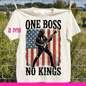 Può includere: T-shirt color crema con una grafica di un chitarrista che suona davanti a una bandiera americana usurata. Il testo "ONE BOSS NO KINGS" è stampato sopra e sotto la grafica in grassetto, in nero. La maglietta è appesa a un filo.
