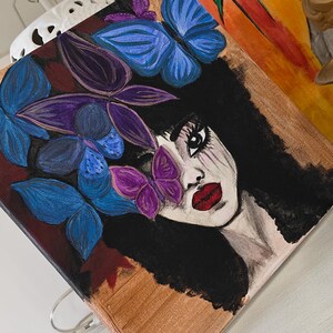 May include: A square canvas painting featuring a woman's face with black hair and red lips. Blue and purple butterflies and leaves adorn her face. The background is a mix of brown and dark red.