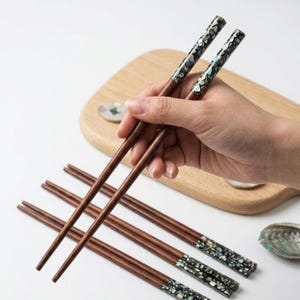 May include: A set of wooden chopsticks with decorative inlaid tips. The chopsticks are brown with a patterned design in shades of black, white, and teal. Several pairs of chopsticks are arranged on a white surface, with one pair being held.