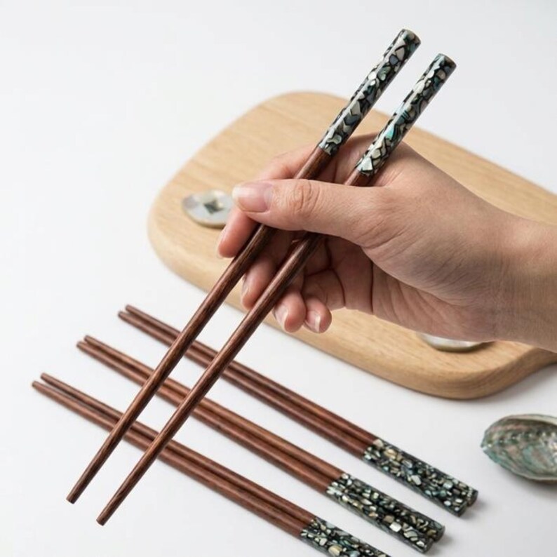 May include: A set of wooden chopsticks with decorative inlaid tips. The chopsticks are brown with a patterned design in shades of black, white, and teal. Several pairs of chopsticks are arranged on a white surface, with one pair being held.