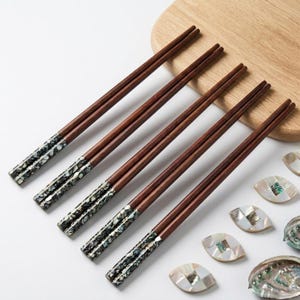 May include: Five pairs of wooden chopsticks with dark brown shafts and decorative, iridescent tips. The chopsticks are arranged diagonally on a white surface, near a wooden cutting board and several decorative shell pieces. A stylish and elegant dining accessory.