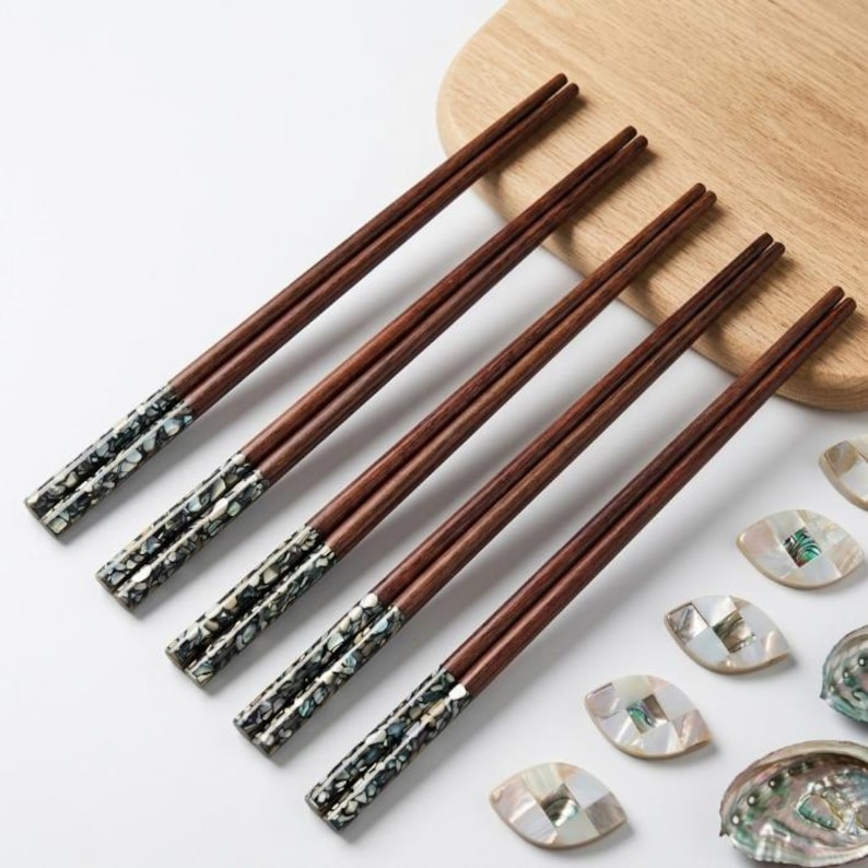 May include: Five pairs of wooden chopsticks with dark brown shafts and decorative, iridescent tips. The chopsticks are arranged diagonally on a white surface, near a wooden cutting board and several decorative shell pieces. A stylish and elegant dining accessory.
