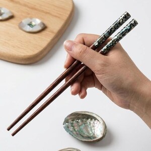 May include: A pair of wooden chopsticks with decorative, iridescent tips are held in a hand. The chopsticks are dark brown, and the tips have a mother-of-pearl design. An abalone shell and wooden cutting board are in the background.