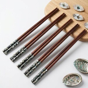May include: Five pairs of wooden chopsticks with decorative mother-of-pearl inlaid tips. The chopsticks are a dark brown color and are arranged diagonally. The tips have a shimmering, iridescent quality. The image also includes decorative shell pieces.