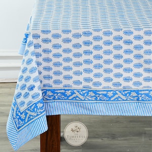 Peut inclure: Une nappe blanche ornée d'un motif répétitif de feuilles bleues sur toute la surface. La bordure présente un dessin horizontal de vignes florales en bleu et blanc, terminé par un bord inférieur à rayures verticales bleues et blanches.