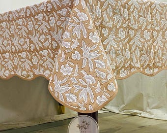 May include: A tan tablecloth featuring a repeating white floral and leaf pattern printed across the fabric. The textile has a scalloped edge finish along the bottom border. A circular logo with the text THE TIMELESS TEXTILE is visible.