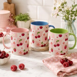 May include: Three ceramic mugs with cherry and floral patterns. The mugs have pink, blue, and green interiors and handles. The mugs are decorated with red cherries, pink flowers, and hearts on a pink and white checkered background.