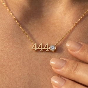 May include: A gold-toned necklace featuring the number 444 in a polished metal finish, accented by a single round, clear, faceted stone set in a circular bezel next to the numbers, suspended from a delicate, thin gold-toned chain.