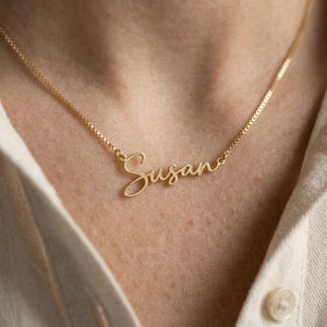 May include: A gold-toned necklace featuring a delicate box chain and a central nameplate pendant in a cursive script font that spells out the name Susan. The metal has a polished finish and the pendant is integrated directly into the chain.