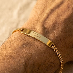 May include: A gold-toned metal chain bracelet featuring a central rectangular identification plate. The plate is engraved with the name Elijah in a clear, dark serif font. The bracelet consists of interlocking curb links that connect to the plate.