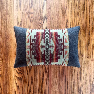 May include: Rectangular decorative pillow with a Southwestern geometric pattern in red, orange, and cream on a light green background. The pillow has dark gray end panels and is set on a wooden surface.