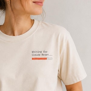 Peut inclure: Un t-shirt à manches courtes de couleur crème avec un petit motif sur la poitrine supérieure gauche. Le motif affiche le texte "Waiting for Claude Reset..." dans une police machine à écrire noire au-dessus d'une barre de chargement orange.